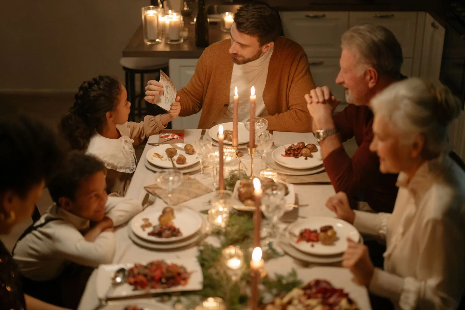 A multi-generational family seated around a dinner table mid-conversation, with one adult leaning in attentively to follow a story