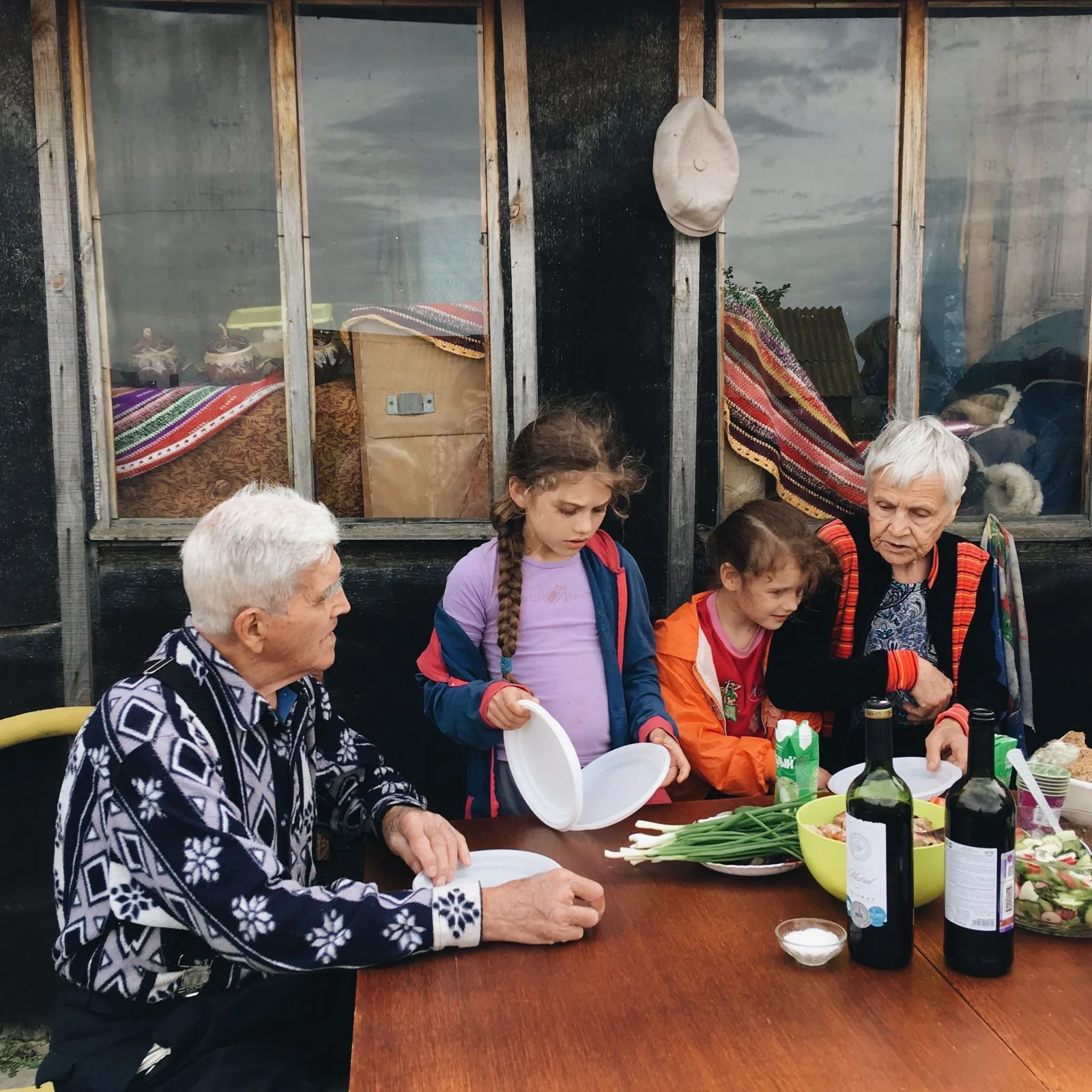 A senior grandparent and grandchildren laughing together while setting a dinner table, illustrating the family connection captioning glasses help preserve for cochlear implant users